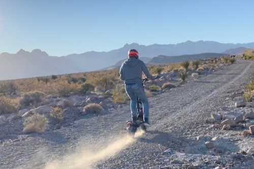 man riding electric scooter in dessert, facing away from camera, dust kicking up